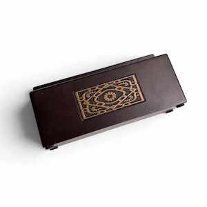 Peut inclure: Une boîte rectangulaire en bois brun foncé avec une incrustation dorée encastrée au centre. L'incrustation présente un motif géométrique symétrique complexe avec des motifs floraux stylisés et des lignes entrelacées sur un fond sombre.