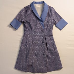 May include: A lavender-colored dress with a floral pattern, featuring a double-breasted design with six buttons. The dress has a light blue collar and cuffs, and short sleeves. The dress is approximately knee-length.