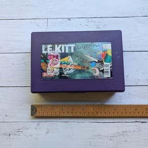 Peut inclure: Une boîte de rangement rectangulaire en plastique violet étiquetée LE KITT avec un graphique montrant des fournitures de couture, notamment des ciseaux, du fil et un crayon. Une règle en bois placée sous la boîte indique une longueur de 28 cm.
