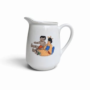 May include: A white ceramic creamer jug featuring a colourful graphic illustration of two figures sitting together. One figure has dark hair and wears a yellow patterned garment, while the other figure has dark hair and wears orange clothing.