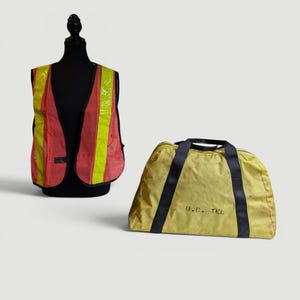 May include: A high-visibility safety vest in red and yellow, displayed on a black mannequin. Beside it is a yellow duffel bag with black straps and the text "B.C. TEL."