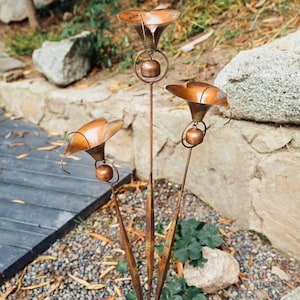 May include: A copper-coloured metal garden stake featuring three flower-shaped bells. Each bell is suspended from a wide, open petal structure attached to a thin vertical rod. The metal has a warm, burnished finish with visible dark patina accents.