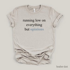Flat lay of a heather dust t-shirt featuring the phrase "running low on everything but opinions" in black and muted dusty blue lettering.