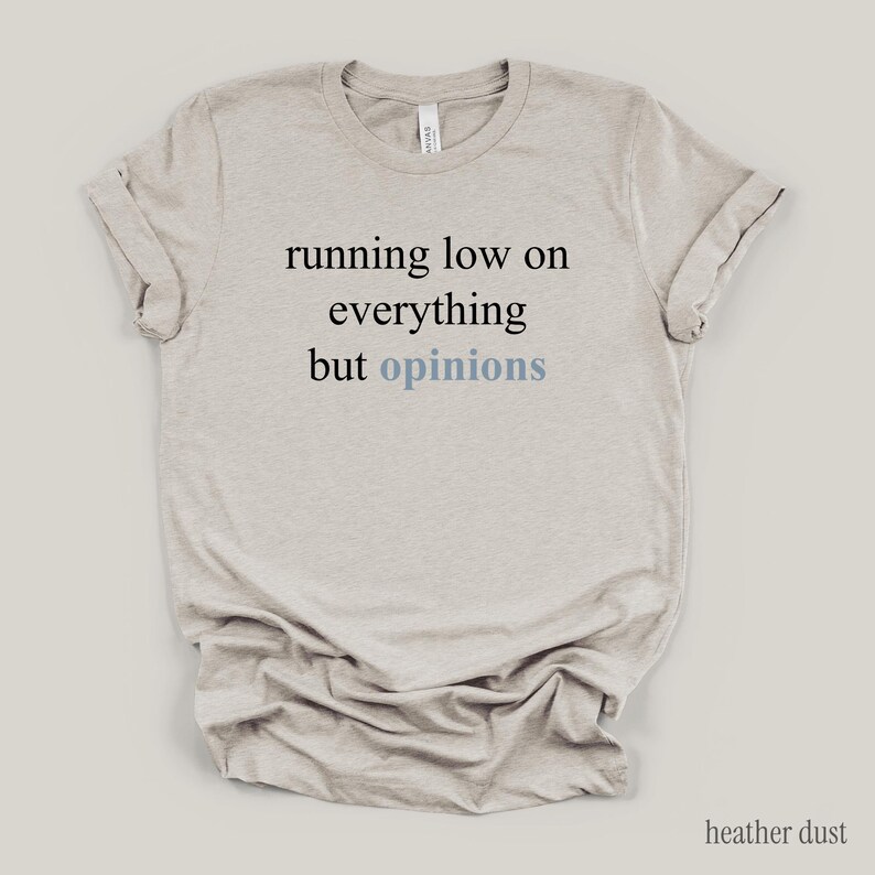 Flat lay of a heather dust t-shirt featuring the phrase "running low on everything but opinions" in black and muted dusty blue lettering.