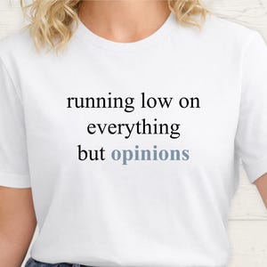 Close up of a model wearing a white t-shirt featuring the phrase "running low on everything but opinions" in black and muted dusty blue lettering.