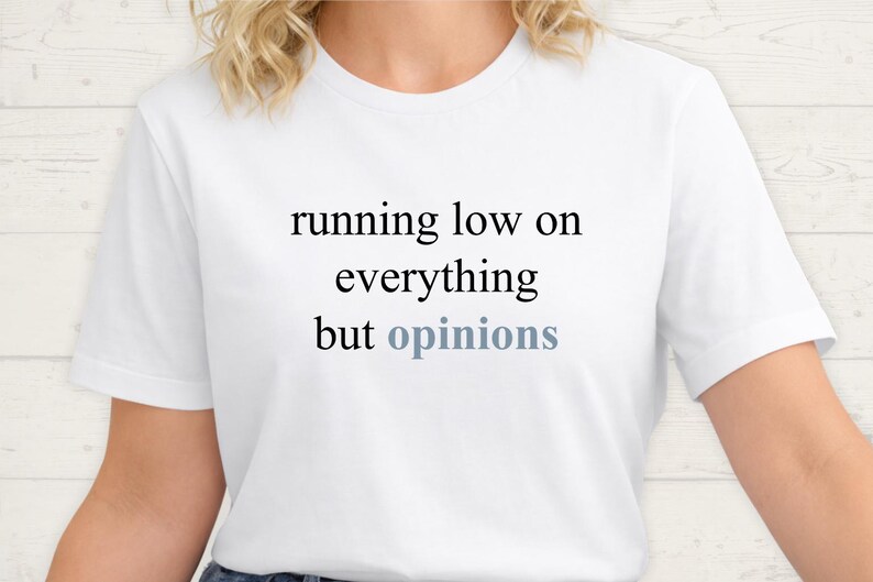 Close up of a model wearing a white t-shirt featuring the phrase "running low on everything but opinions" in black and muted dusty blue lettering.