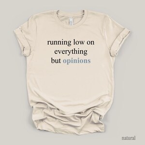 Flat lay of a cream t-shirt featuring the phrase "running low on everything but opinions" in black and muted dusty blue lettering.
