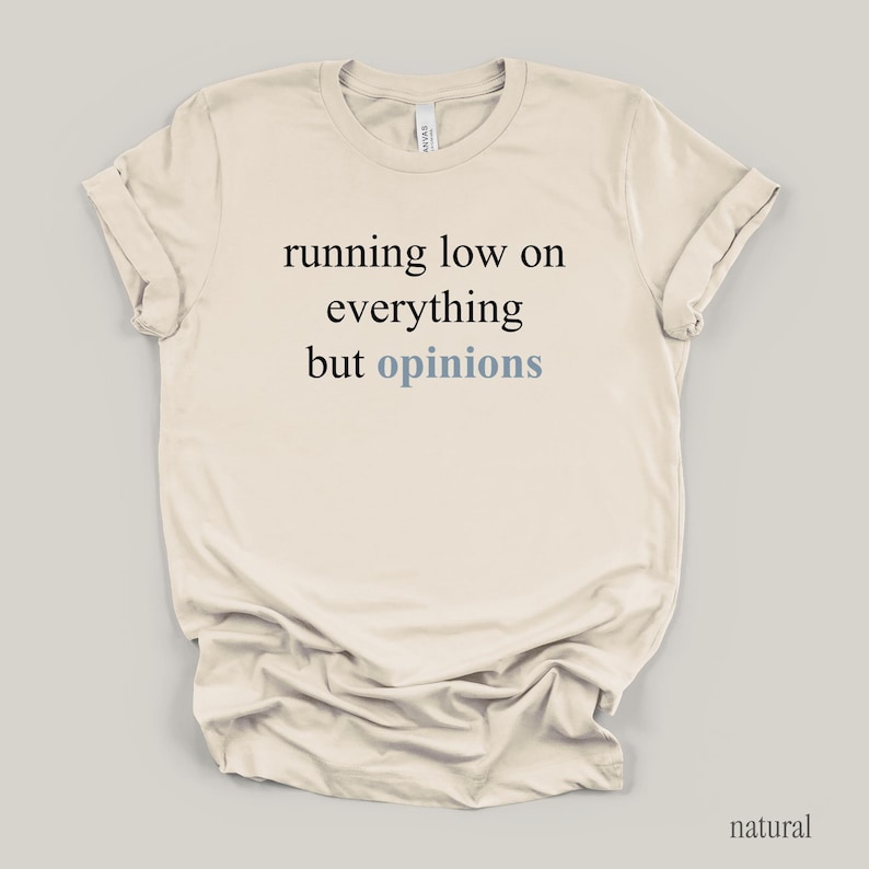 Flat lay of a cream t-shirt featuring the phrase "running low on everything but opinions" in black and muted dusty blue lettering.