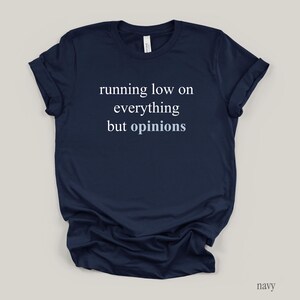 Flat lay of a navy t-shirt featuring the phrase "running low on everything but opinions" in white and muted dusty blue lettering.