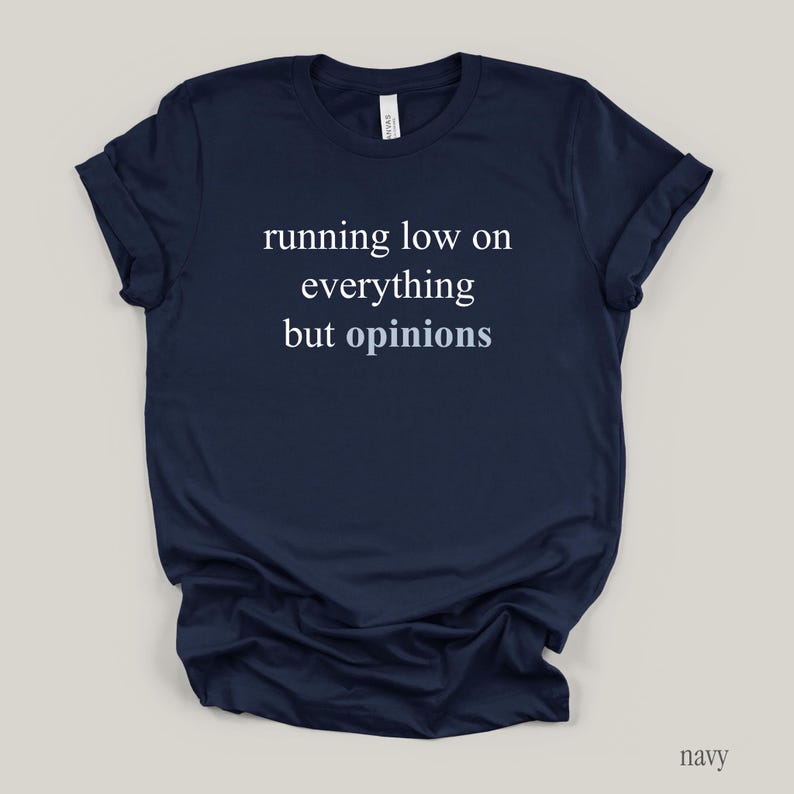 Flat lay of a navy t-shirt featuring the phrase "running low on everything but opinions" in white and muted dusty blue lettering.