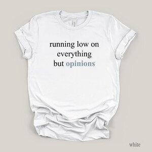 Flat lay of a white t-shirt featuring the phrase "running low on everything but opinions" in black and muted dusty blue lettering.