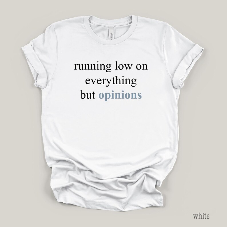 Flat lay of a white t-shirt featuring the phrase "running low on everything but opinions" in black and muted dusty blue lettering.