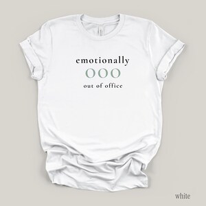 Flat lay of a white t-shirt featuring the phrase "emotionally ooo out of office" in black and sage lettering.