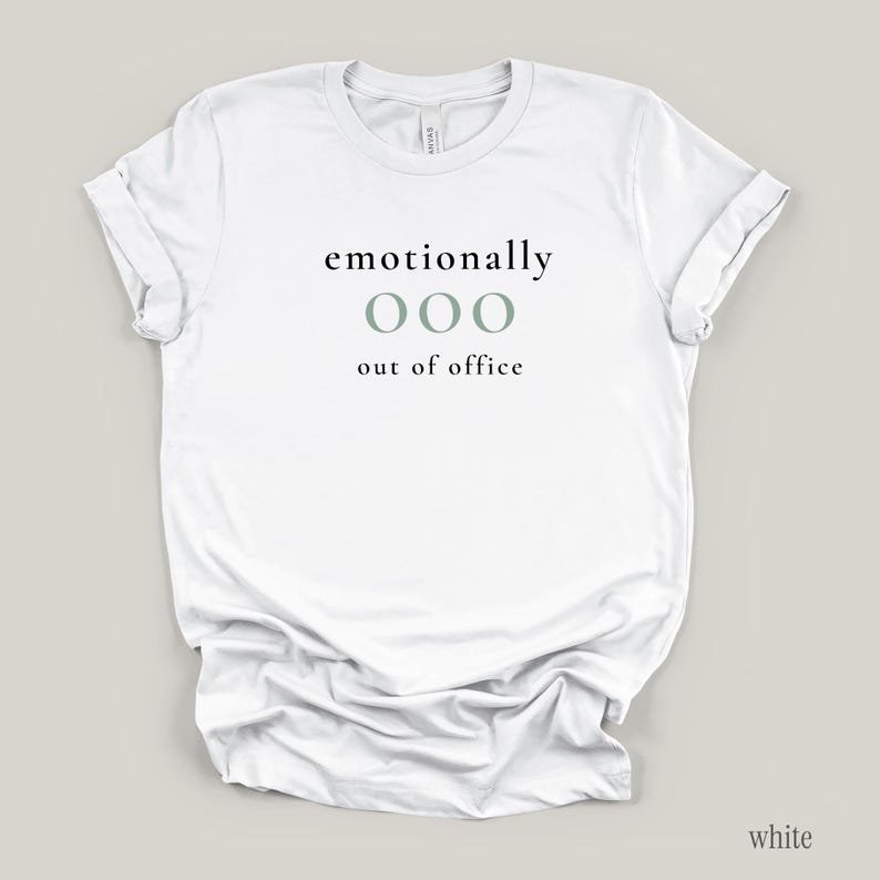 Flat lay of a white t-shirt featuring the phrase "emotionally ooo out of office" in black and sage lettering.