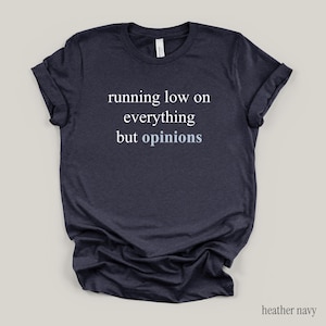 Flat lay of a heather navy t-shirt featuring the phrase "running low on everything but opinions" in white and muted dusty blue lettering.