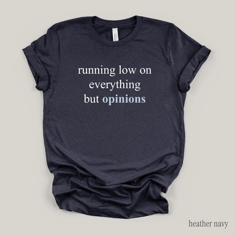 Flat lay of a heather navy t-shirt featuring the phrase "running low on everything but opinions" in white and muted dusty blue lettering.