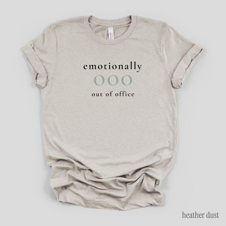 Flat lay of a heather dust t-shirt featuring the phrase "emotionally ooo out of office" in black and sage lettering.