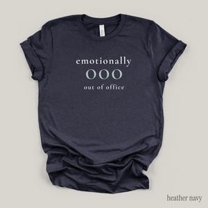 Flat lay of a heather navy t-shirt featuring the phrase "emotionally ooo out of office" in white and sage lettering.