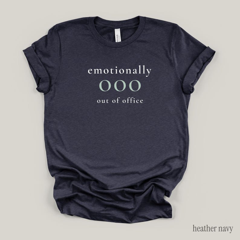 Flat lay of a heather navy t-shirt featuring the phrase "emotionally ooo out of office" in white and sage lettering.