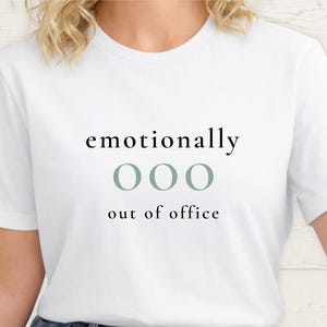Close up of a model wearing a white t-shirt featuring the phrase "emotionally ooo out of office" in black and sage lettering.