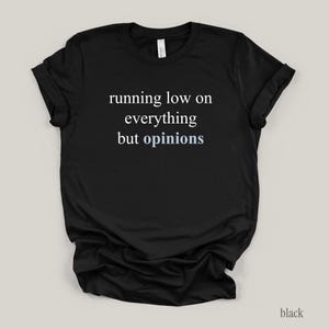 Flat lay of a black t-shirt featuring the phrase "running low on everything but opinions" in white and muted dusty blue lettering.