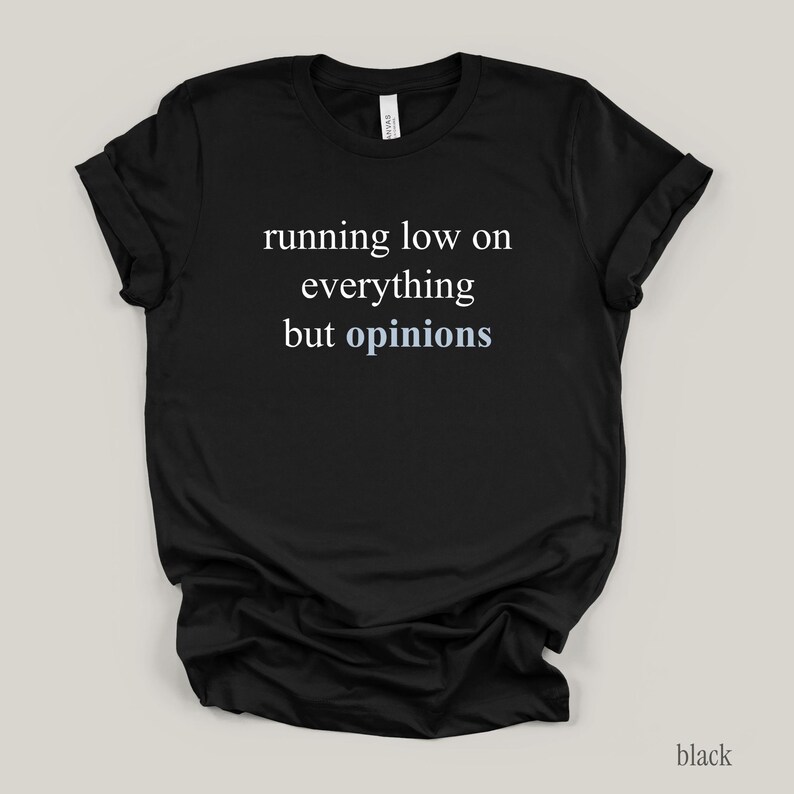 Flat lay of a black t-shirt featuring the phrase "running low on everything but opinions" in white and muted dusty blue lettering.