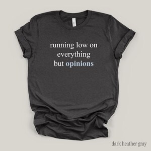 Flat lay of a dark gray t-shirt featuring the phrase "running low on everything but opinions" in white and muted dusty blue lettering.