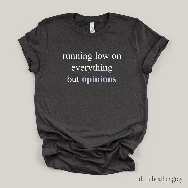 Flat lay of a dark gray t-shirt featuring the phrase "running low on everything but opinions" in white and muted dusty blue lettering.