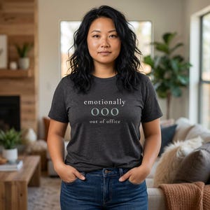 Model wearing a dark gray t-shirt featuring the phrase "emotionally ooo out of office" in white and sage lettering.