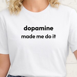 Puede incluir: Camiseta blanca con la frase negra "dopamine made me do it" impresa en el centro. La camiseta tiene mangas cortas y cuello redondo. La persona lleva la camiseta.