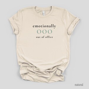 Flat lay of a cream t-shirt featuring the phrase "emotionally ooo out of office" in black and sage lettering.