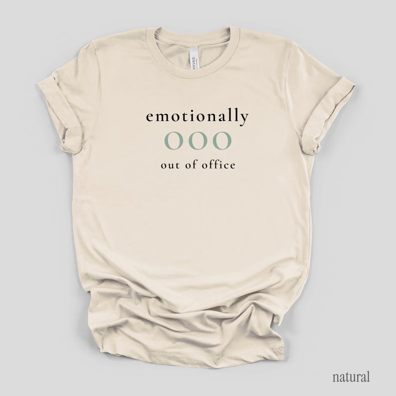 Flat lay of a cream t-shirt featuring the phrase "emotionally ooo out of office" in black and sage lettering.