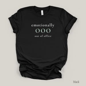 Flat lay of a black t-shirt featuring the phrase "emotionally ooo out of office" in white and sage lettering.