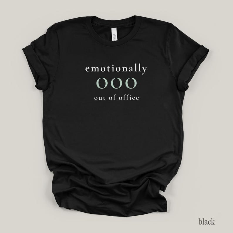 Flat lay of a black t-shirt featuring the phrase "emotionally ooo out of office" in white and sage lettering.
