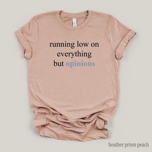 Flat lay of a light peach t-shirt featuring the phrase "running low on everything but opinions" in black and muted dusty blue lettering.