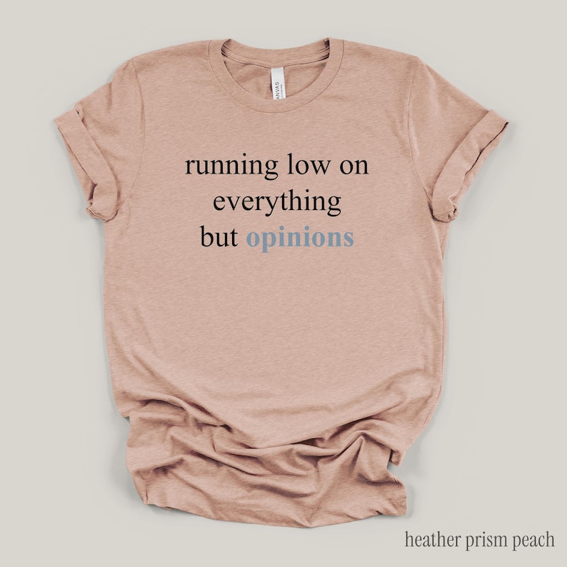 Flat lay of a light peach t-shirt featuring the phrase "running low on everything but opinions" in black and muted dusty blue lettering.