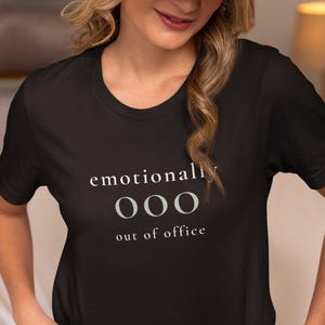 Model wearing a black t-shirt featuring the phrase "emotionally ooo out of office" in white and sage lettering.