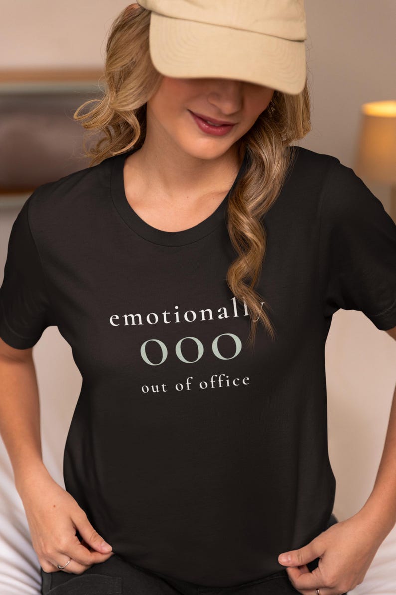 Model wearing a black t-shirt featuring the phrase "emotionally ooo out of office" in white and sage lettering.