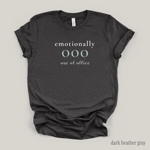 Flat lay of a dark gray t-shirt featuring the phrase "emotionally ooo out of office" in white and sage lettering.