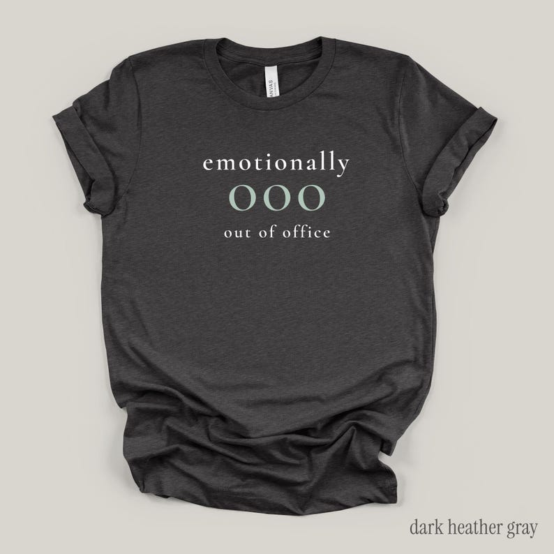 Flat lay of a dark gray t-shirt featuring the phrase "emotionally ooo out of office" in white and sage lettering.