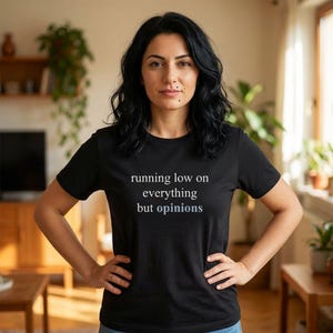 Model wearing a black t-shirt featuring the phrase "running low on everything but opinions" in white and muted dusty blue lettering.