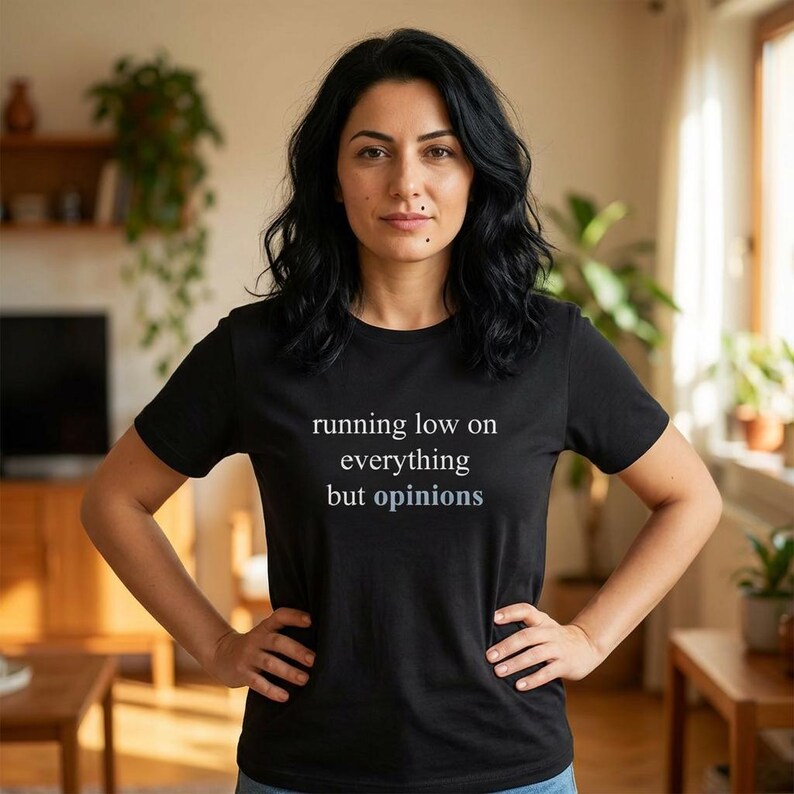 Model wearing a black t-shirt featuring the phrase "running low on everything but opinions" in white and muted dusty blue lettering.