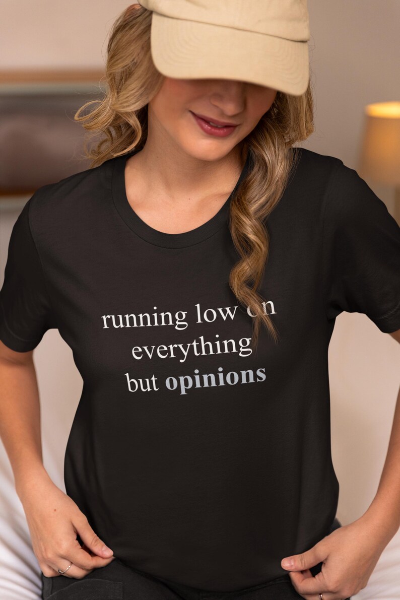 Model wearing a black t-shirt featuring the phrase "running low on everything but opinions" in white and muted dusty blue lettering.