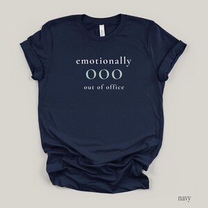 Flat lay of a navy t-shirt featuring the phrase "emotionally ooo out of office" in white and sage lettering.