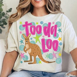 Può includere: T-shirt color crema con un canguro dei cartoni animati e la scritta rosa "Tooda Loo". Il design include bolle blu e stelle colorate. Una maglietta grafica divertente e giocosa.