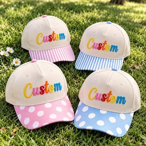 Puede incluir: Cuatro gorras de béisbol con coronas de color crema y la palabra Custom bordada en una fuente degradada multicolor. Dos gorras tienen viseras a rayas en rosa y azul, mientras que las otras dos tienen viseras de lunares en rosa y azul.