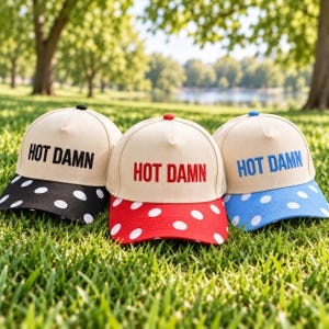May include: Three baseball caps with beige crowns and the text HOT DAMN embroidered on the front. The left cap has a black brim with white polka dots, the center cap has a red brim with white polka dots, and the right cap has a blue brim with white polka dots.