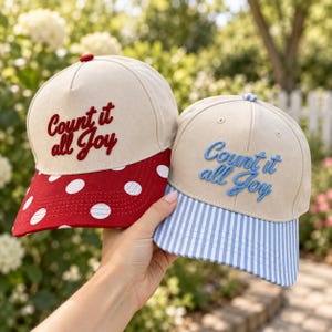May include: Two baseball caps with cream-colored crowns and embroidered text reading Count it all Joy. One cap features a red brim with white polka dots, while the other has a light blue and white striped brim. Both caps have a top button.