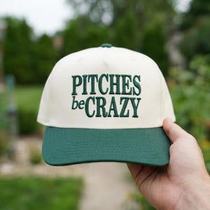 Op de afbeelding: Een crèmekleurige baseballpet met een donkergroene klep en donkergroene geborduurde tekst op de voorkant die PITCHES beCRAZY leest. De pet heeft een gestructureerde kroon en een platte klep, met een tweekleurig ontwerp en opvallende letters.