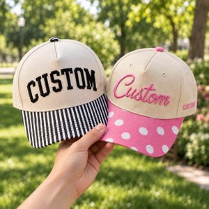 Puede incluir: Dos gorras de béisbol con coronas beige. La gorra izquierda tiene el texto bordado en negro CUSTOM y una visera a rayas blancas y negras. La gorra derecha tiene el texto bordado en rosa Custom y una visera rosa con lunares blancos.